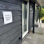 Dark wooden house exterior with a white sign reading 'ZONNETJE WIJNTJE GENIET' mounted beside a glass door on the porch area.