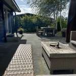 Sunlit backyard patio with wicker sofa, matching coffee table, and a dining table set among trees.