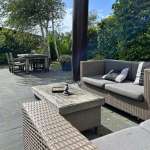 Patio seating area with gray wicker sofa and matching coffee table on stone tiles; a wooden dining table and chairs in the background.