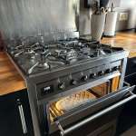 Stainless steel gas range with six burners; oven door open showing a round pizza inside; wooden countertops and knife block in the background.