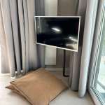 Flat-screen TV on a stand beside a large brown floor pillow, with gray curtains and a glass door nearby.